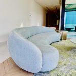 “Curved to perfection – this sofa goals in ice-blue bouclé.” A luxurious, organic-shaped curved sofa in soft ice-blue/gray plush upholstery positioned in a bright modern living space with herringbone wood flooring, layered neutral rugs, and large windows letting in abundant natural light.