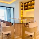“When your dining table looks like a piece of modern art you can actually eat at.” A luxurious modern dining room with a large oval glass-top table on a fluted natural wood pedestal base, surrounded by eight sculptural cream-upholstered dining chairs with curved wooden frames. Warm ambient lighting from recessed ceiling LEDs and a backlit onyx shelving unit creates an elegant glow.