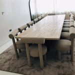 “That moment you realize the chairs are comfier than most sofas.” A sleek modern dining room with a long rectangular travertine stone table surrounded by ten plush taupe bouclé “Fat” style chairs with cylindrical wrapped backrests and chunky rounded legs. The set sits on a large neutral area rug under bright natural light from tall curtained windows, with a minimalist arched floor mirror in the background.