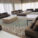 “When your living room can comfortably host the whole family… and the neighbors.” A grand, brightly lit formal majlis with multiple low taupe upholstered sectional sofas arranged in a large open square around a central travertine coffee table. Traditional Persian-style rugs in soft turquoise and cream tones layer the glossy white marble floor, while floor-to-ceiling sheer white curtains diffuse abundant natural light.