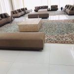 “When your living room can host the entire family reunion and still look this calm.” A spacious, brightly lit formal majlis/living room with multiple low, plush taupe-brown modular sofas arranged in symmetrical clusters around large travertine coffee tables. An ornate traditional Persian-style area rug in soft blues and creams anchors the central seating area, set against glossy white marble floors and floor-to-ceiling sheer curtains.