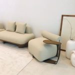 One chair to make the whole room feel like a permanent hug A minimalist modern living corner featuring a plush cream bouclé lounge chair with an integrated cylindrical bolster and a matching low sectional sofa. A sculptural bronze-finish C-shaped side table slides under the chair. Large white ceramic vases with dried branches and a simple framed artwork complete the serene neutral scene.