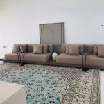 Modern majlis minimalism at its finest – low, long, and impossibly chic A minimalist modern majlis with a long, low built-in taupe upholstered sectional running wall-to-wall, accented with matching throw pillows. Two sleek black metal C-shaped side tables slide over the seating. A large oriental rug in soft turquoise and cream lies in front, under a contemporary beige geometric wall artwork.