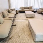 “When your furniture looks this soft, shoes are officially banned.” A spacious, minimalist living room furnished entirely in soft cream and beige tones, featuring ultra-modern curved modular sofas, oversized organic-shaped ottomans, and sleek side tables. Floor-to-ceiling sheer white curtains diffuse bright natural light across layered neutral rugs.