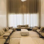 “Low seating + high ceilings + endless beige = pure luxury serenity.” A grand, double-height minimalist majlis-style living room with floor-to-ceiling sheer white curtains and heavy taupe drapes framing tall windows. Low, plush beige modular sofas form a large U-shaped seating arrangement around oversized cream ottomans on layered neutral rugs, creating a serene, luxurious conversation pit.