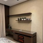 A minimalist console vignette blending warm wood tones, neutral textures, and curated decorative accents. Wooden console table with open shelves, red cushions, black vases, potted plants, and beige textured wall.
