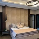 Vertical elegance and warm tones define this luxurious bedroom retreat. Bedroom with tall upholstered headboard, beige and brown bedding, pendant lights, and circular chandelier.