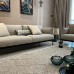 A serene palette and sculptural accents bring elegance and tranquility to this contemporary lounge. Living room with beige sofas, abstract blue and gold artwork, glass vases, and sheer curtains.