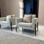 Minimalist design meets cozy elegance in this modern seating arrangement. Interior with two beige armchairs, black metal legs, textured pillows, tiled floor, and natural lighting.