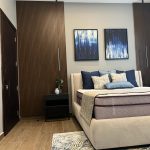 A clean palette and geometric accents bring serenity and style to this modern bedroom layout. Bedroom with beige upholstered bed, blue and gray pillows, wood panel wall, abstract paintings, and black nightstands.