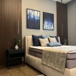 A soothing palette and sculptural accents bring balance and elegance to this modern bedroom design. Bedroom with beige upholstered bed, blue and gray pillows, circular rug, wood slat wall, and abstract art.