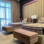 Soft hues and layered textures create a serene and luxurious twin bedroom arrangement. Bedroom with two twin beds, pink benches, beige headboard, and floor-to-ceiling curtains.