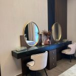 Symmetry and soft tones elevate this modern vanity into a refined space for beauty and reflection. Elegant vanity setup with black countertop, gold-framed mirrors, pink chairs, and decorative accessories.