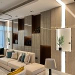 Layered textures and curated lighting bring warmth and elegance to this contemporary lounge design. Living room with beige sectional sofa, colorful throw pillows, wood-paneled wall, built-in shelves, and chandelier.