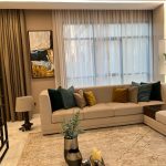 Natural light and rich accents bring warmth and elegance to this contemporary living space. Living room with beige sectional sofa, green and mustard pillows, wooden coffee table, and abstract wall art.