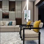 A refined mix of textures and tones brings warmth and sophistication to this modern lounge setup. Living room with beige sofa, patterned armchairs, teal and mustard cushions, wood-paneled wall, and decorative shelving.