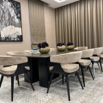 A neutral palette and sculptural decor bring balance and elegance to this modern dining space. Dining room with black table, ten beige upholstered chairs, golden floral centerpieces, and abstract wall art.