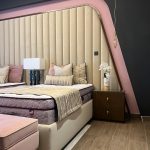 A sculptural bedroom composition blending warm neutrals, soft textures, and artistic wall geometry. Beige upholstered bed with mauve mattress, paneled headboard, pink accent wall, and gold-accented nightstand.